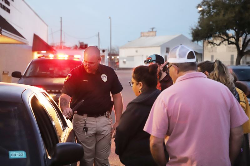 In a parking lot, a uniformed police officer shines a light into a car while a small group watches