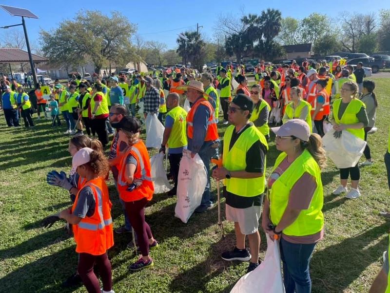 Large group of people in a park wearing orange and yellow work vests with trash bags and grabbers