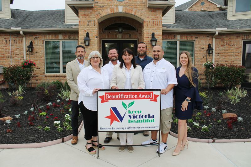 Group photo with KVB sign in front of orange brick apartment office with flowerbeds