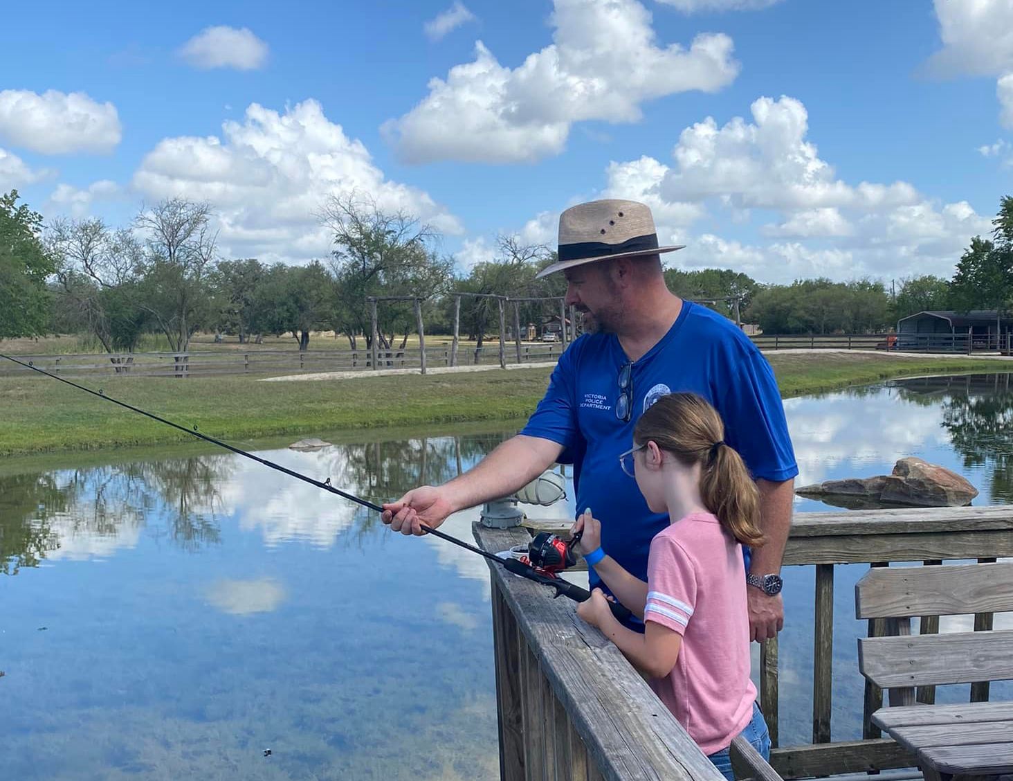 A man in a police department shirt helps a girl hold a fishing rod