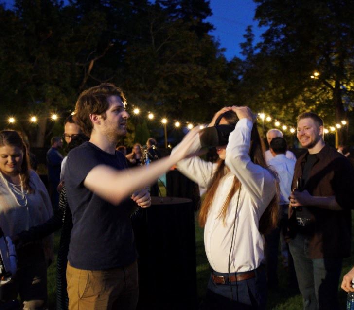 In an outdoor area lit with hanging lights, a man helps a woman put on a virtual reality headset.