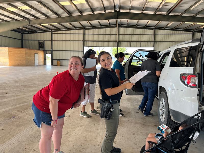 Two women, one holding a clipboard, smile and pose for a photo outside a car
