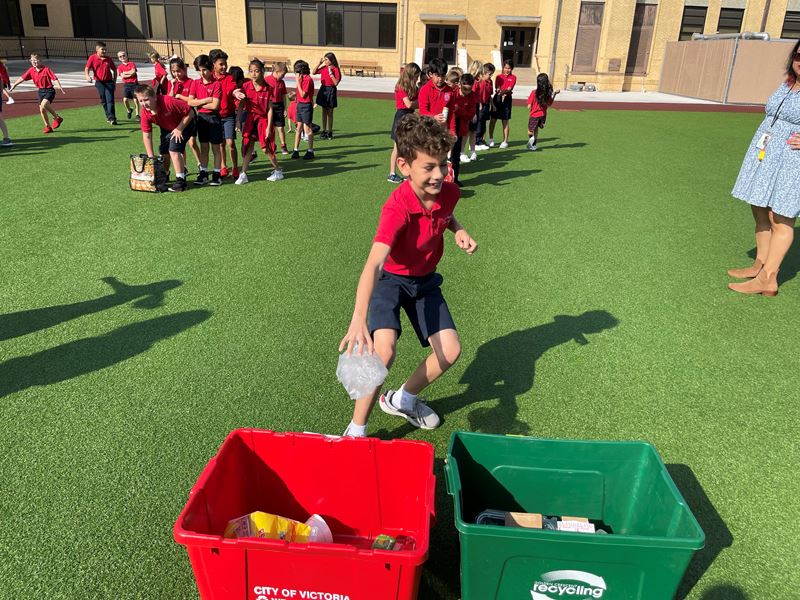 A boy in a red shirt runs to put trash in a bin. Other children in red shirts wait in lines.