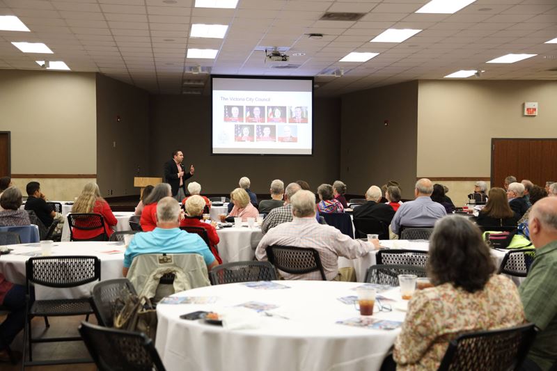 Large group of people seated at round tables watch a presentation by City Manager Jesús Garza.