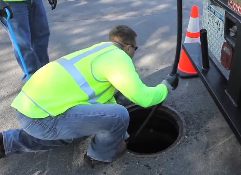 A person wearing a yellow safety vest inserts a long, narrow hose into a sewer through a manhole