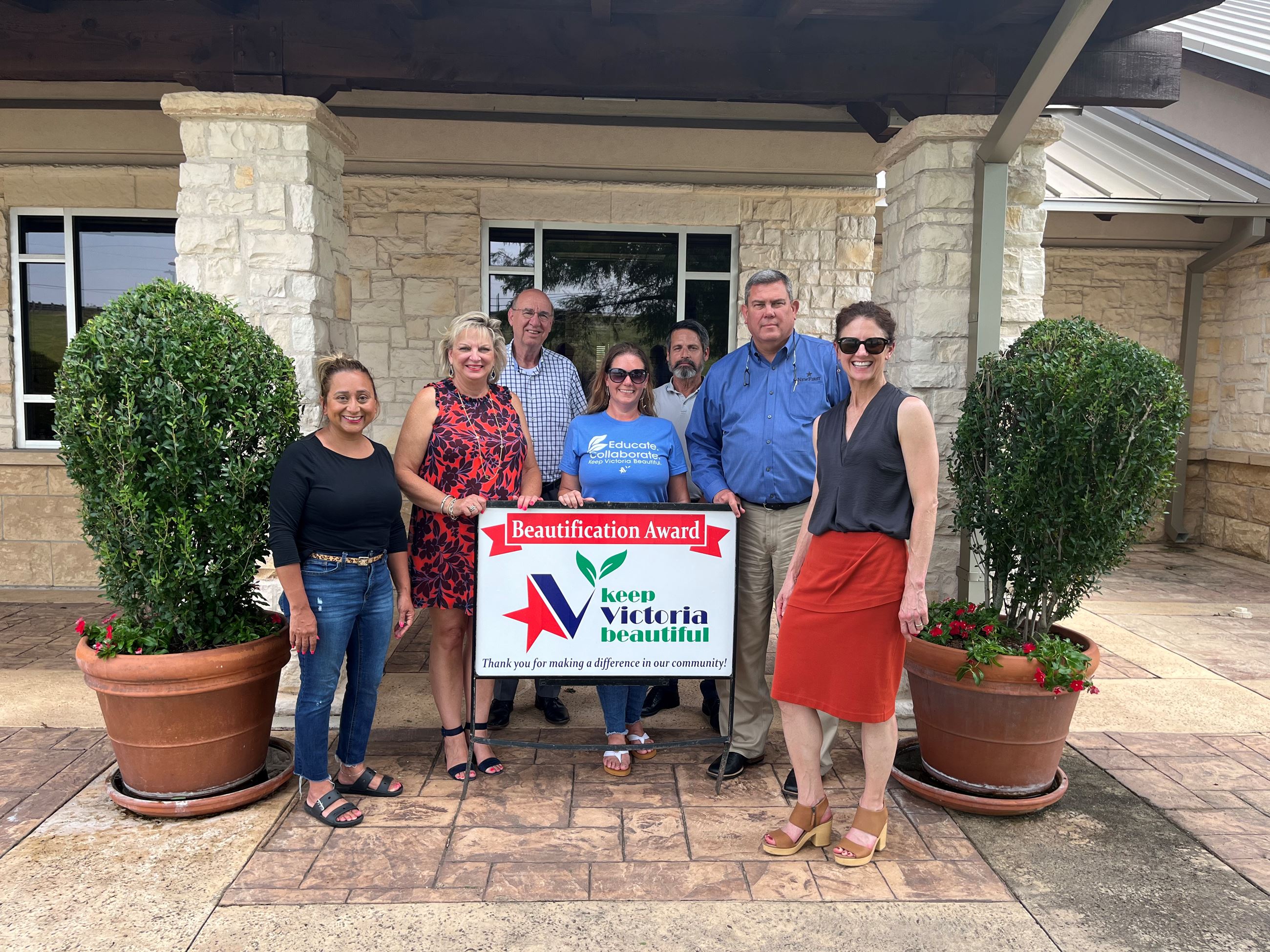 Group photo with KVB sign in front of two potted ficuses surrounded by flowers