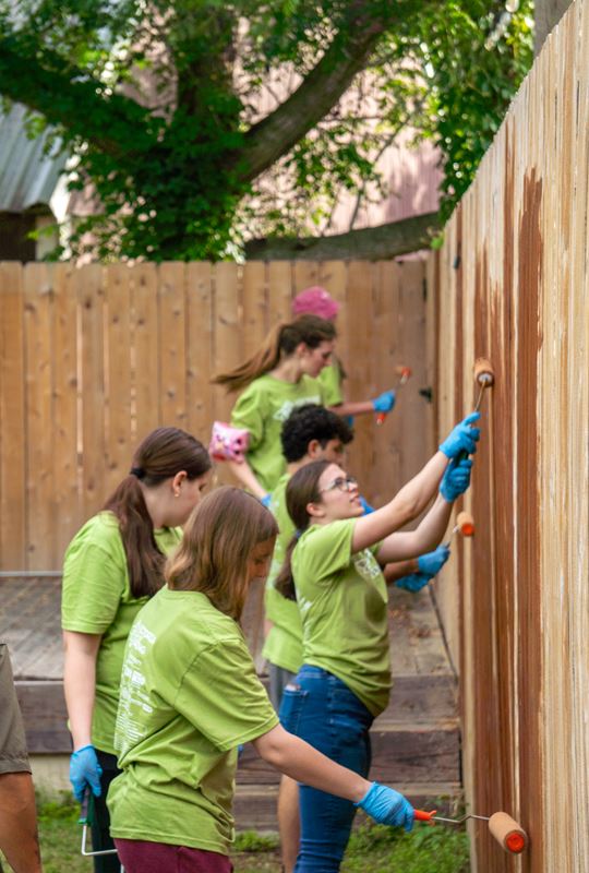 Teens in matching green T-shirts paint a high wooden fence brown.