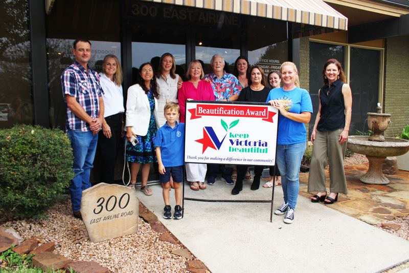 Group photo with KVB sign in front of building with a bed of shrubs and pebbles and a water fountain