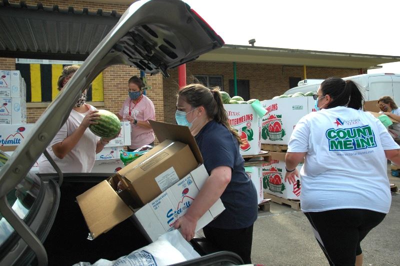 People in masks load food into a car trunk. Boxes of food are stacked in the background.