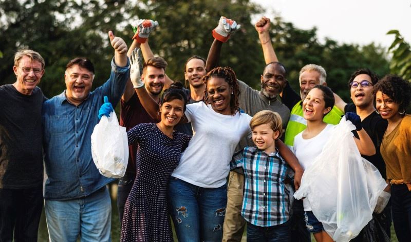 Group of people outdoors smiling with trash bags and gloves. Some raise hands or give thumbs-up.