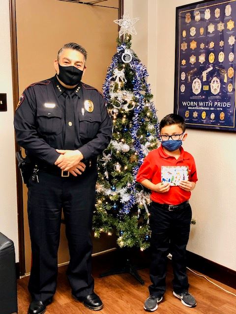 Policeman in uniform stands next to child holding card. Both stand in front of a Christmas tree.