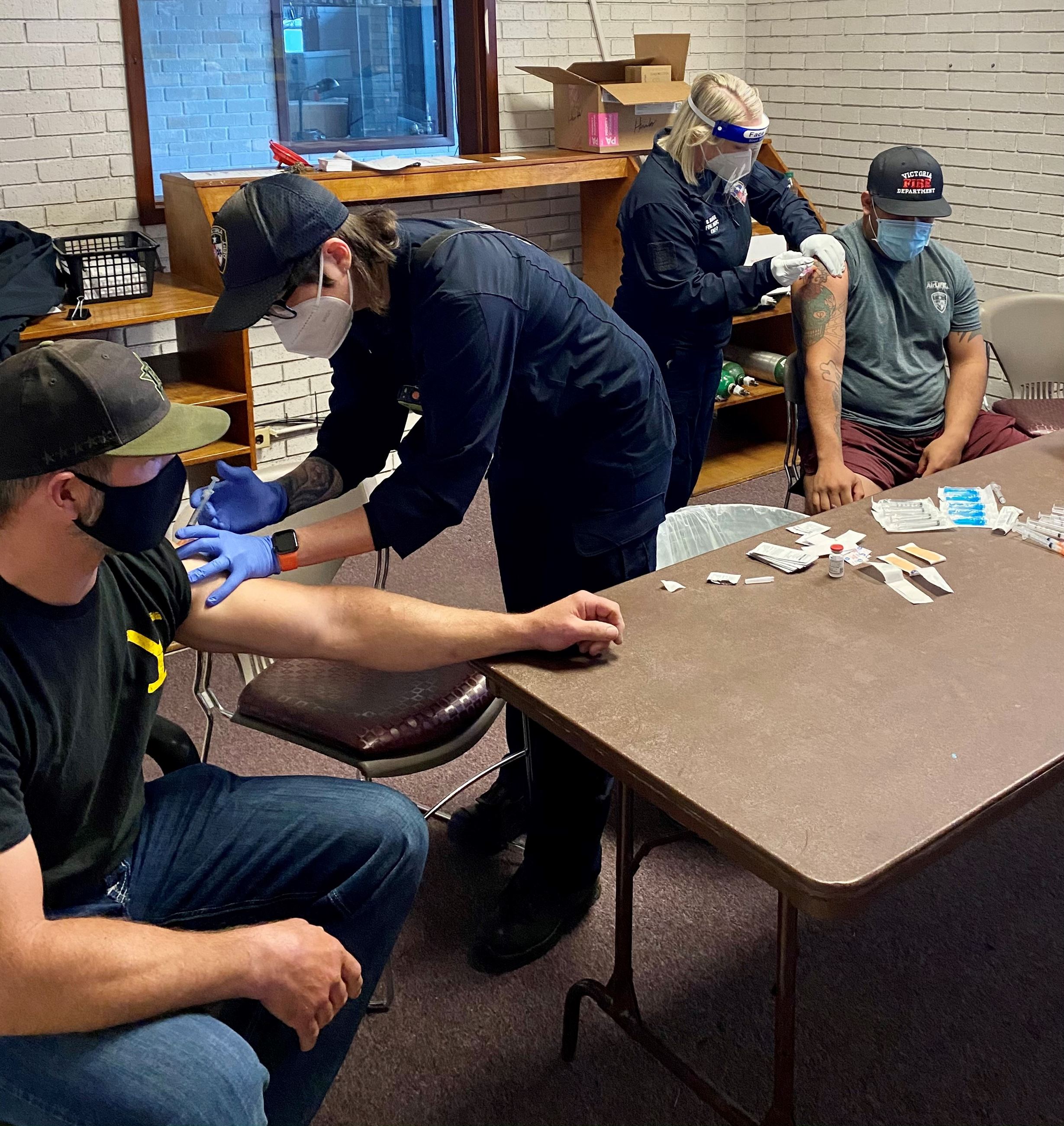 Two female fire department employees administer injections to two male fire department employees.