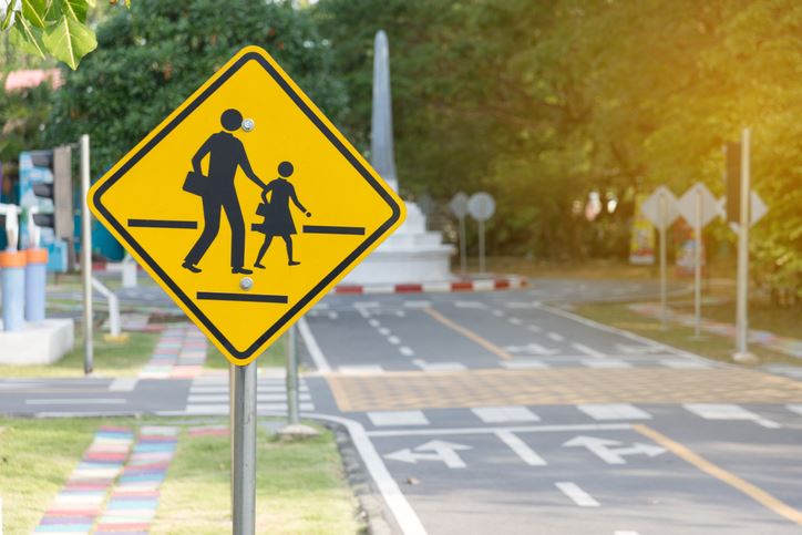 A crossing sign featuring an adult and child near a street marked with crosswalks and turn lanes.