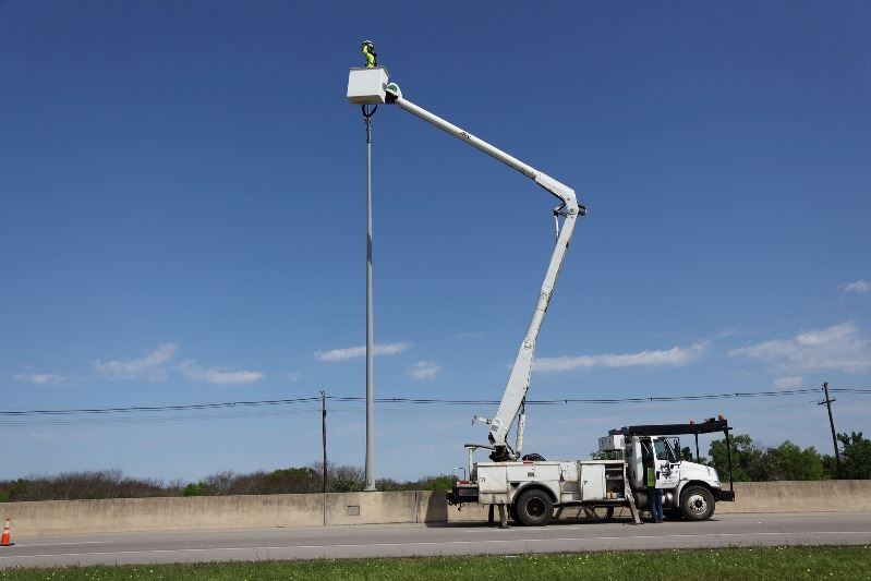 A cherry picker style truck extends its basket up to a street light on a highway.