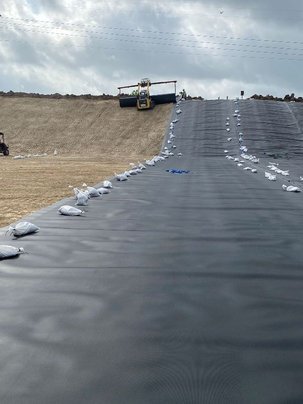 A rolling machine lays down a black plastic liner on a wide, flat indented area