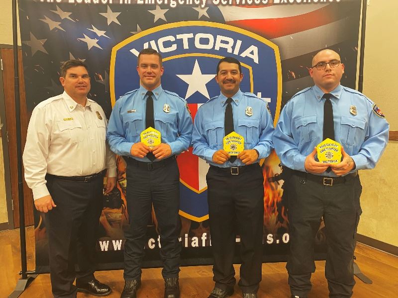 Four people in firefighter dress clothes. Three have blue shirts and hold yellow patches.