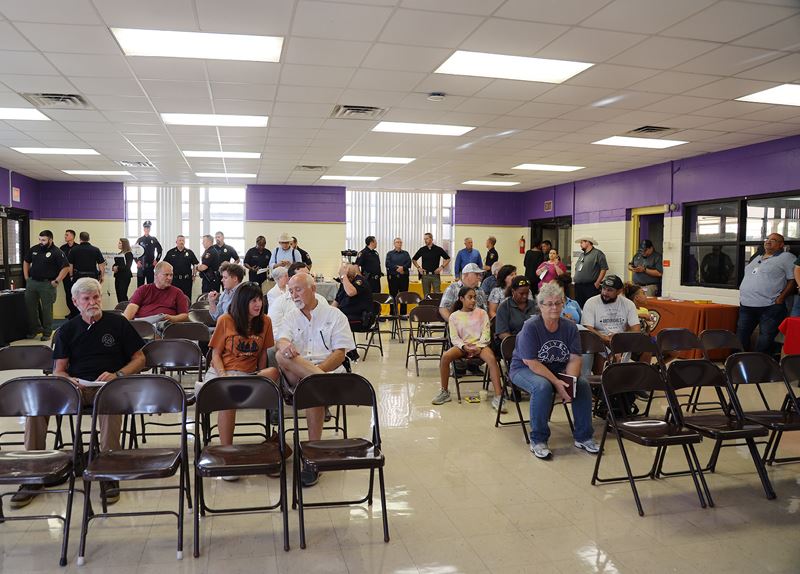 People sit in rows of folding chairs and talk amongst themselves. Police officers stand in the back.