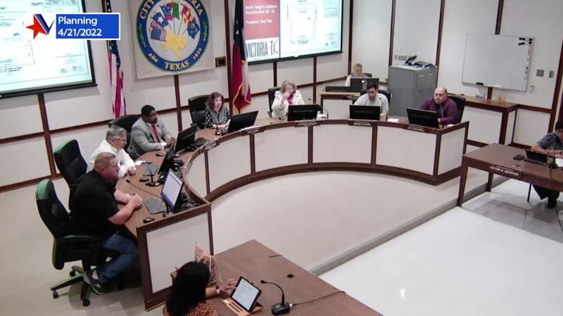 People seated at council dais. Projector screens in the back show a slide show.
