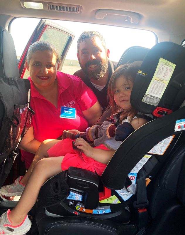 A child in a car seat. Two adults stand in the car doorway.