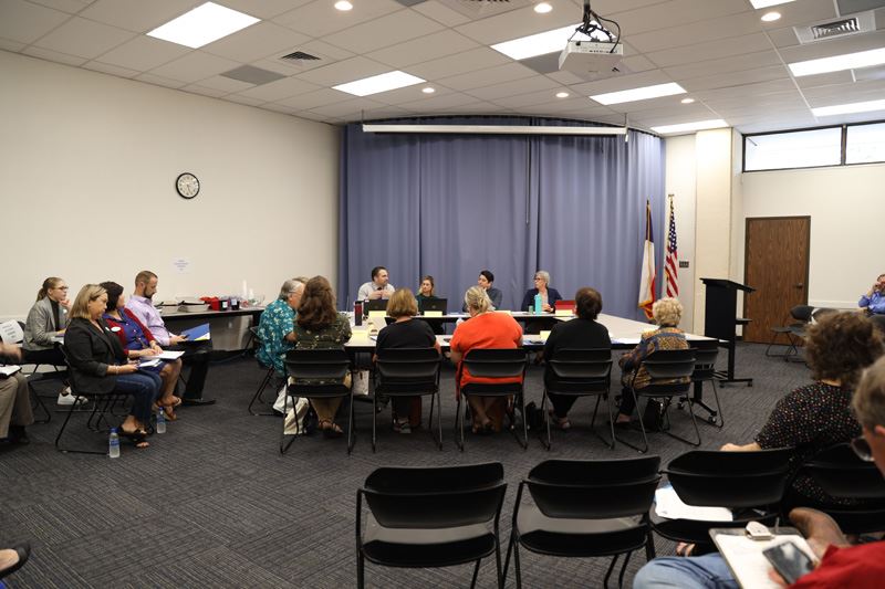People seated at rectangle tables arranged in a square. Other people sit in rows of folding chairs.