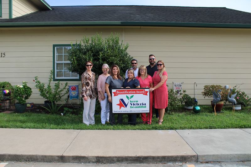 Group photo with KVB sign in front of a building with a row of shrubs, small trees and lawn decor