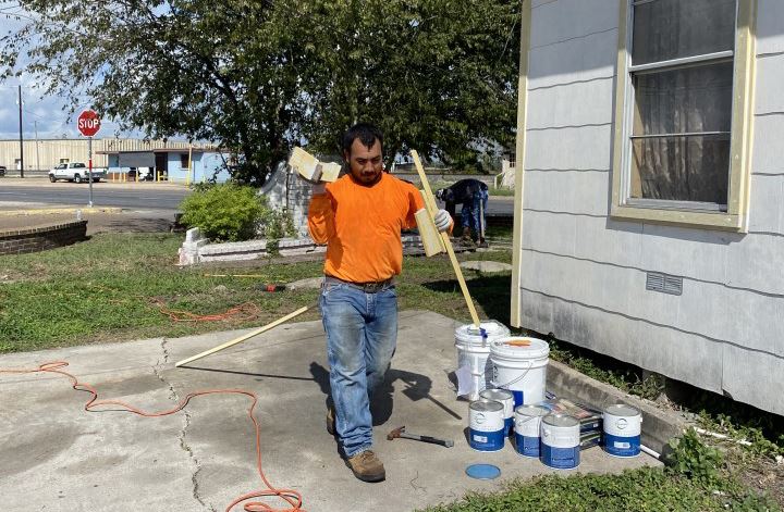 A man in an orange shirt holds construction materials in both hands near a house.
