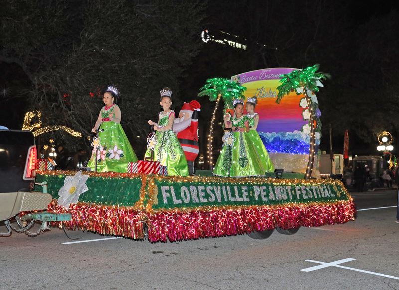 Parade float with a tropical scene with many Christmas lights and four girls in green dresses Opens in new window