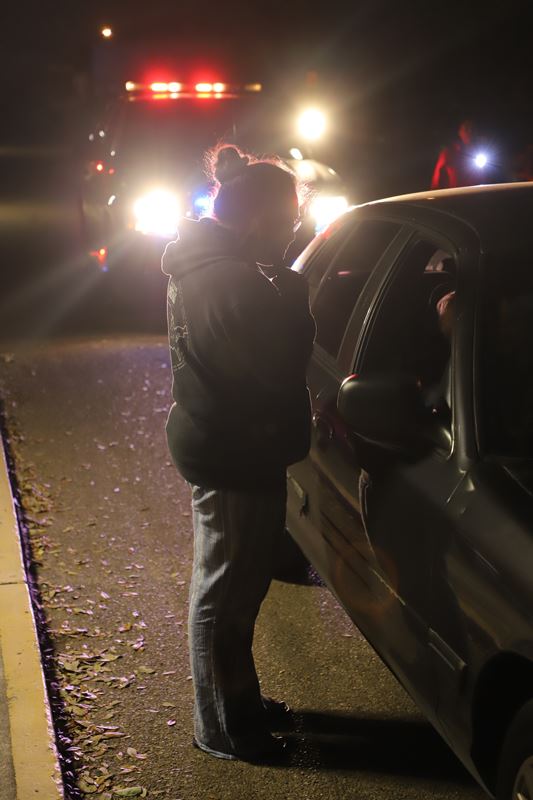 In the dark, a woman stands next to the passenger window of a car. Police car lights on behind.