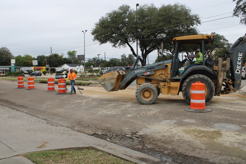 Road construction. A scooper moves dirt, a man sweeps and another man walks dragging a shovel.