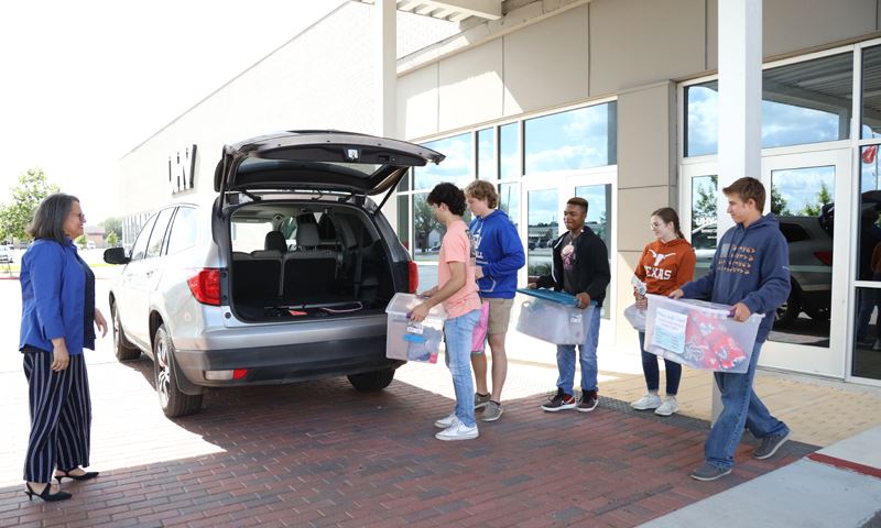 Students carry plastic tubs and bags to the open back of a silver van. A woman stands nearby.
