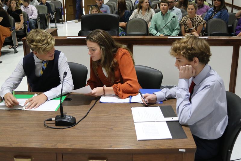 Three teenagers sit around a table in the Municipal Court courtroom with folders in front of them.