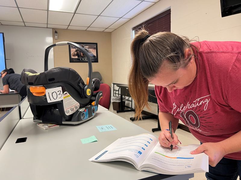A woman writes in a workbook. Near her, a child car seat sits on a table.