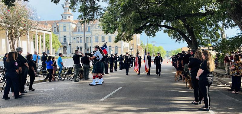 Uniformed police officers line a street downtown. Some march with flags. Musicians play bagpipes.