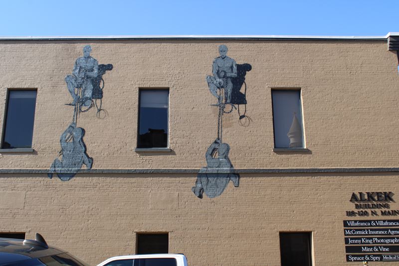 Mural on the side of a building featuring gray human figures helping each other climb