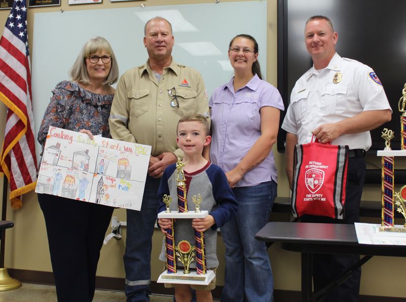 A boy holding a trophy. Four adults stand behind him holding a poster and a red bag.