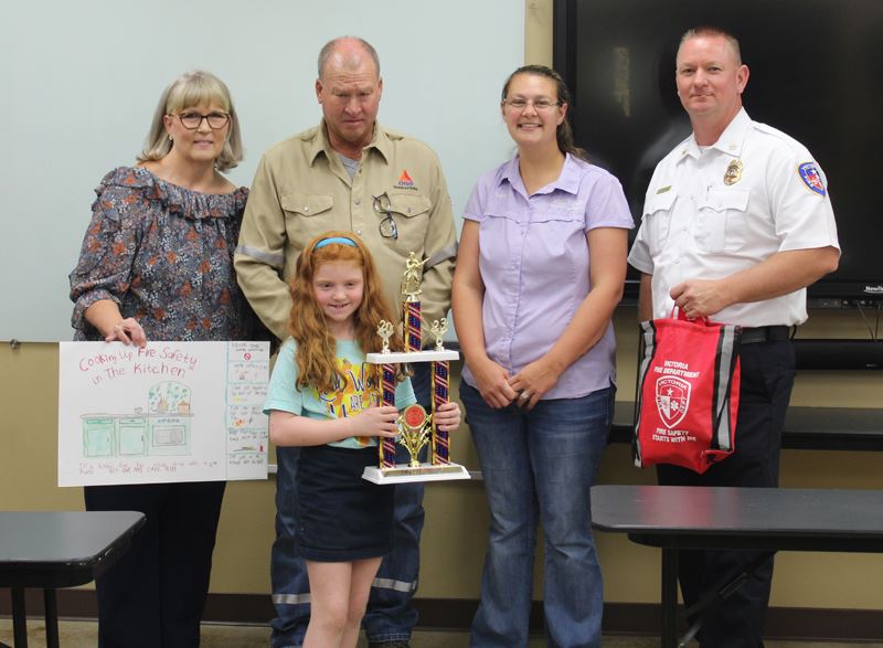 A girl holding a trophy. Four adults stand behind her holding a poster and a red bag.