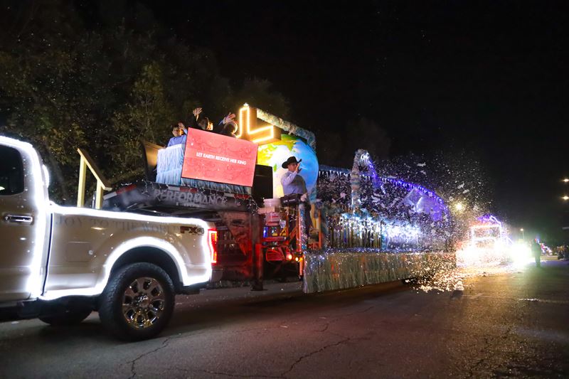Lighted parade float pulled by a lighted truck with a lighted cross and TV playing Joy to the World