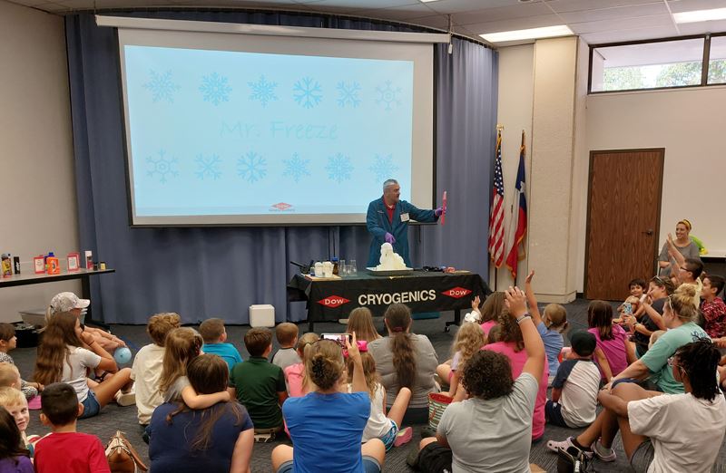 Children sit around a table where a man uses science tools to produce a foaming chemical reaction