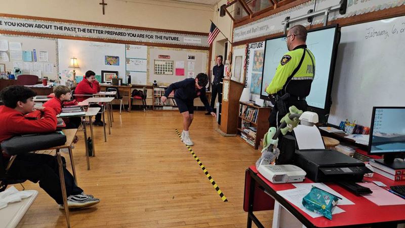 police officer watches as a kid walks on a line in front of a class of children