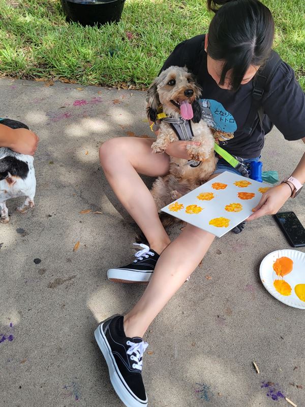 A young woman holds a dog in her lap and uses its paw to make yellow paw prints on paper