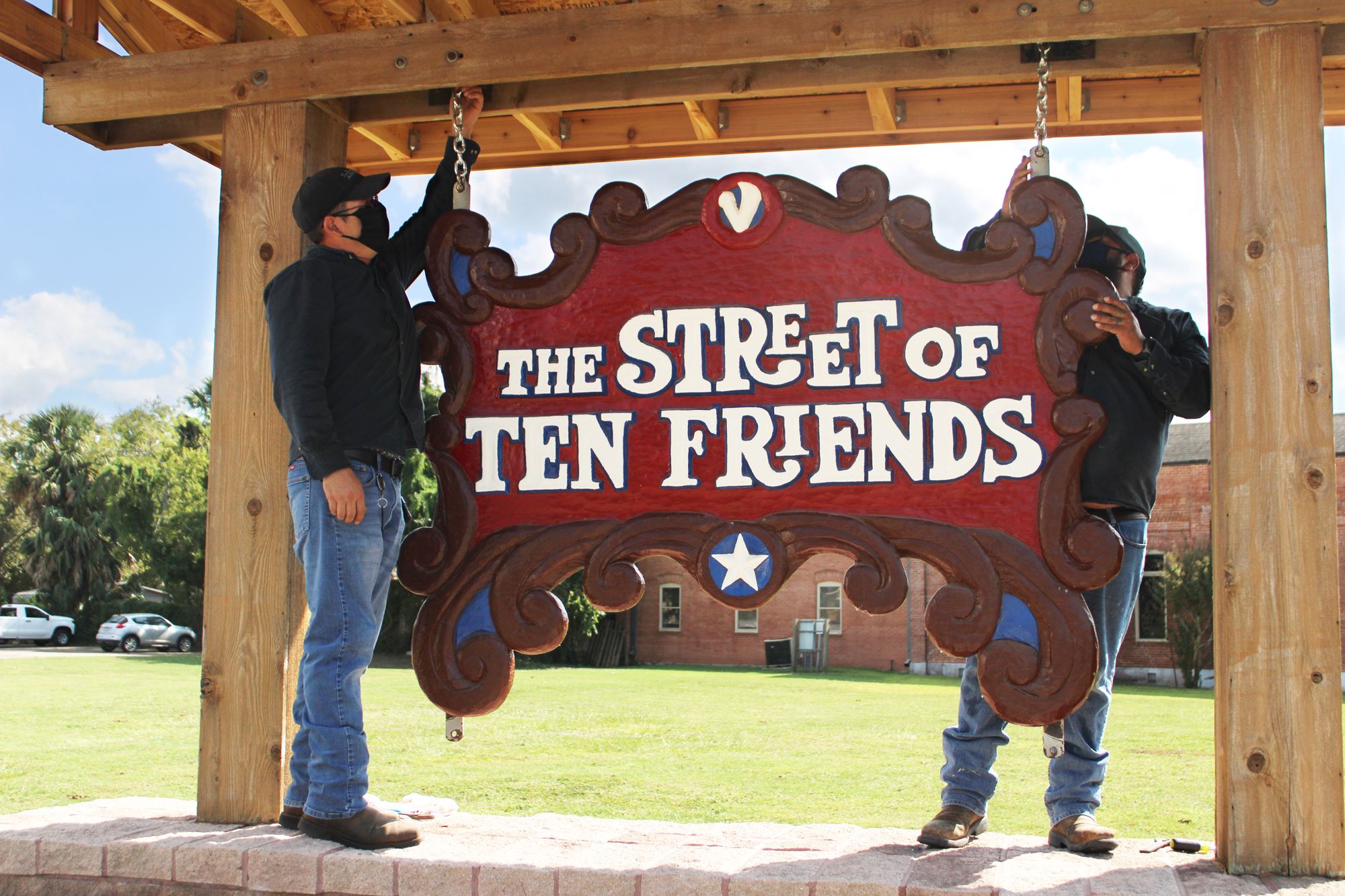 2 men wearing face masks hang a wooden sign under a wooden shelter