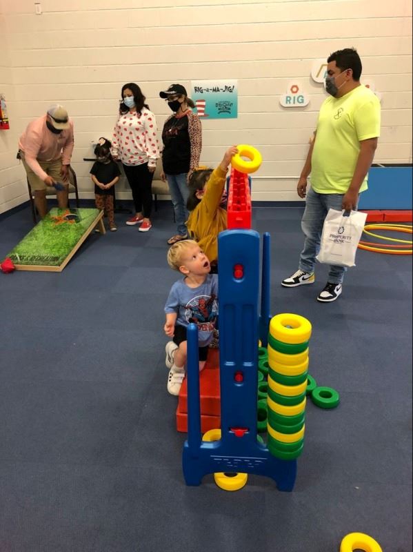 Young kids play with a giant Connect Four set indoors. Hula hoops and other toys in the background.