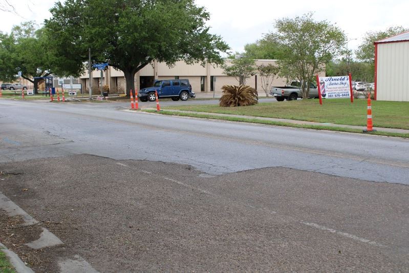 A cracked, patchy road. Traffic cones sit on the grass along the curb.