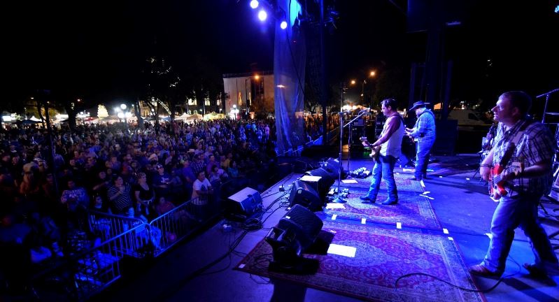 Musicians perform onstage at night outside with blue lights in front of cheering crowd