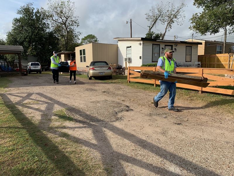 Three people carry bulky trash items down a residential dirt driveway.