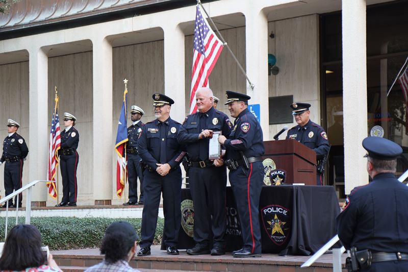 Three uniformed police officers stand at a makeshift stage at the police station. One holds a plaque