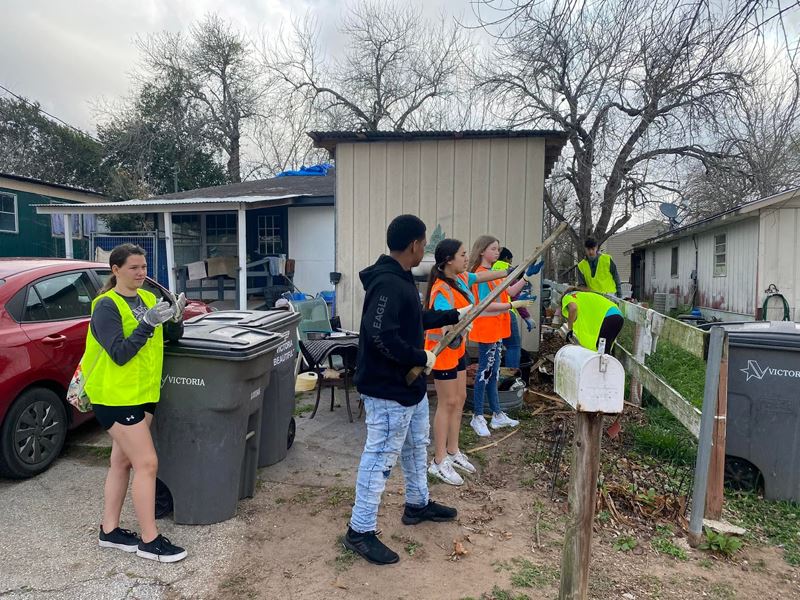 Kids wearing safety vests pick up trash outside a home.