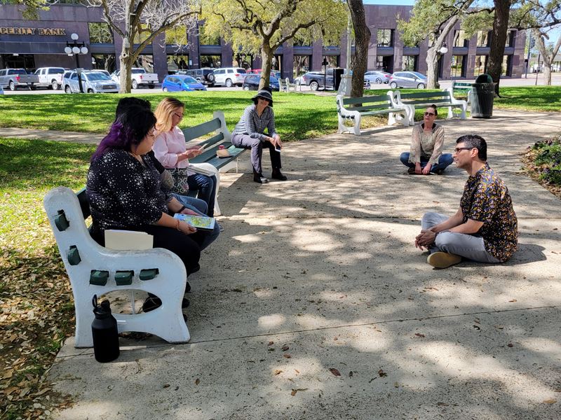 People sit in benches and on the sidewalk at DeLeon Plaza, looking at a seated woman who is reading.