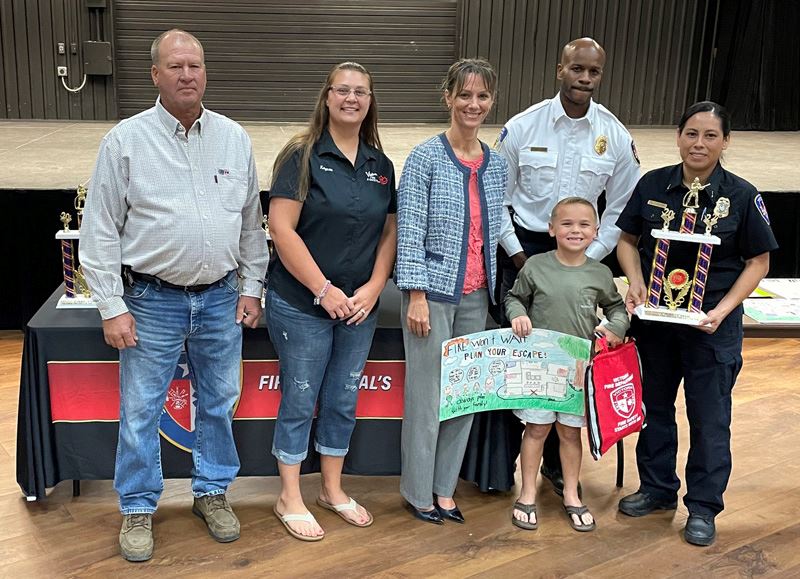 Five adults stand with a child who is holding a hand-drawn poster with fire safety images.